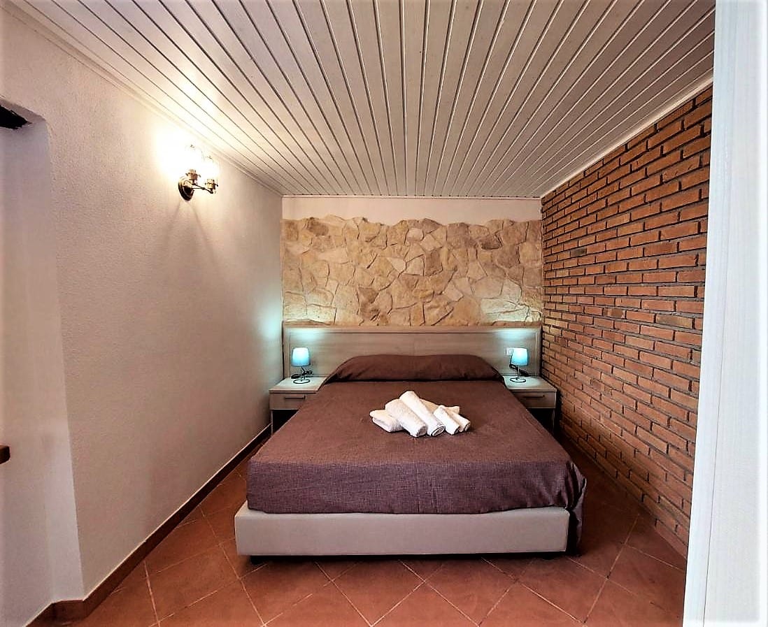 Camera con letto matrimoniale, parete in mattoni e finestra con tende chiare